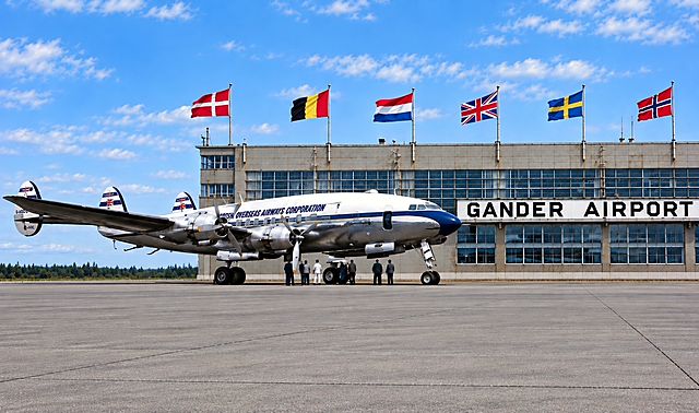 A silver airliner parked in front of the Gander Airport terminal, with the flags of many nations flying above the building